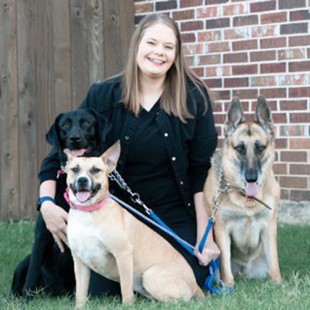 Woman smiling outdoors with three dogs, including a black lab, tan mixed breed, and German shepherd.