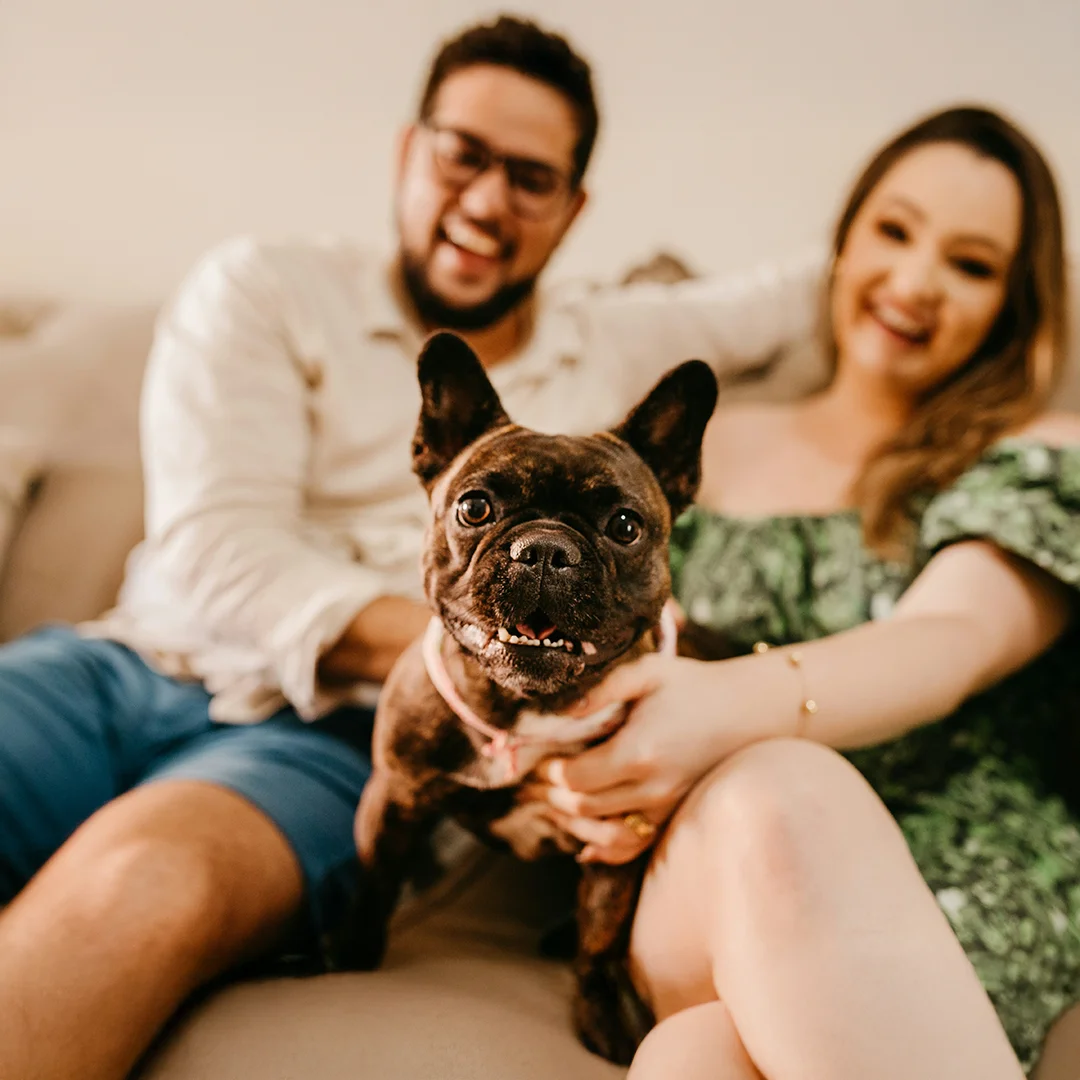 Smiling couple sitting on a couch with a French Bulldog in the foreground looking at the camera.