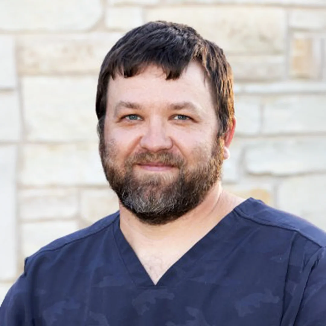 Bearded man in navy scrubs smiling in front of a light stone wall.